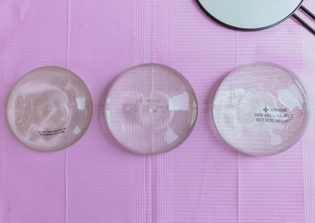 Close up of 3 breast implants lying on a pink surface beside a handheld mirror.